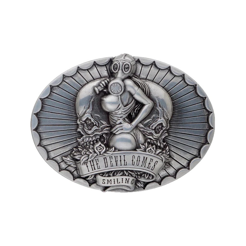 A detailed silver belt buckle with a gothic or biker-inspired design. This fashion accessory features a woman in a gas mask, a large skull, and a banner that reads 'THE DEVIL COMES SMILING'.